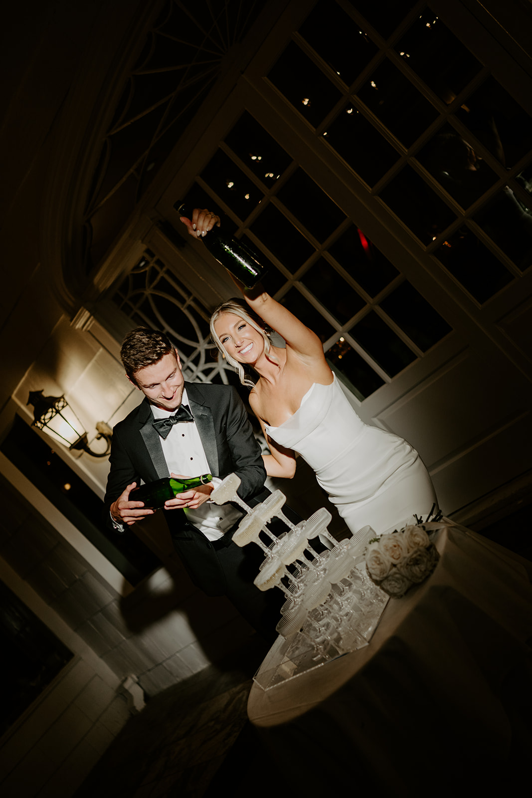 Smiling bride and groom pour champagne into a glass tower at night, celebrating their wedding.